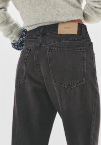 Personne portant un jean en denim noir avec des poches arrière visibles et une étiquette Pull&Bear beige, associé à un pull gris clair.