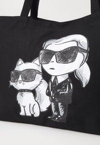 Fekete vászontáska, amelyen egy macska és napszemüveget viselő figura karikatúra terve látható, fehér és szürke színben, textúrált megjelenéssel.