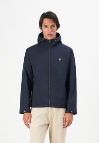 Man wearing a navy hooded jacket with a yellow bird logo on the chest, paired with light beige pants, standing against a white background.
