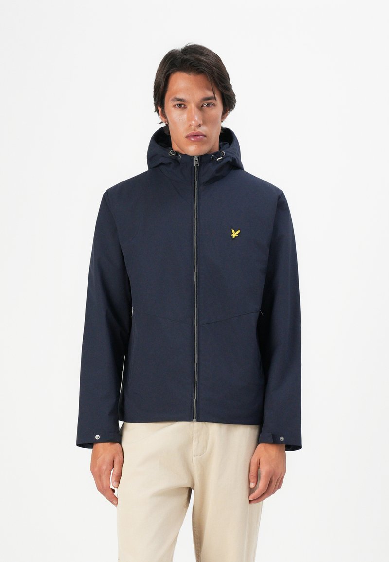 Man wearing a navy hooded jacket with a yellow bird logo on the chest, paired with light beige pants, standing against a white background.