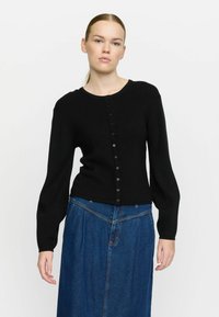 Mustat ribbed-kudotut neuletakit, joissa on pitkät hihat, pyöreä kaula-aukko ja nappisuljenta edessä. Yhdistetty siniseen farkkuhameeseen.