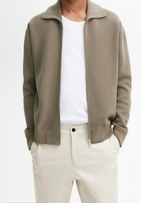 Homme portant une veste zippée taupe sur un t-shirt blanc et un pantalon crème, avec les mains dans les poches, devant un fond blanc uni.