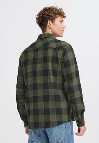 Camicia in flanella a scacchi verdi e neri con maniche lunghe, colletto e polsini con bottoni. Tessuto morbido e orlo curvo nella parte inferiore.