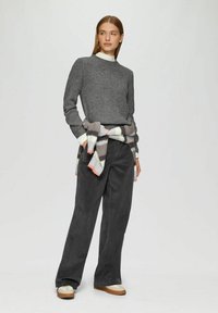 Grauer gerippter Pullover mit hohem Kragen, kombiniert mit dunkelgrauen weiten Hosen. Ein mehrfarbiger karierter Schal ist um die Taille gebunden. Beigefarbene Sneakers.