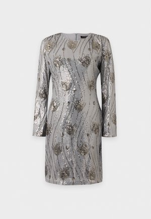 Un mannequin se tient devant un fond blanc, vêtu d’une robe gris argenté à manches longues ornée de sequins à motifs floraux, et de chaussures à talons à brides.