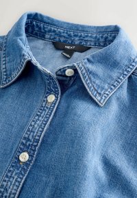 Lyseblå denimskjorte med spids krave, der har to hvide knapper og synlige sømdetaljer. Mærket siger "NEXT."