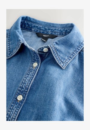 Camisa de mezclilla azul clara con cuello puntiagudo, que presenta dos botones blancos y detalles de costura visibles. La etiqueta dice "NEXT".