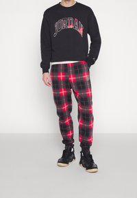 Sudadera negra con la palabra "JORDAN" en letras rojas y blancas, combinada con pantalones de plaid rojos y negros que incluyen un parche de logo y puños elásticos. Zapatillas negras.