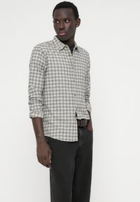 Vyras vilkintis šviesiai pilką ir juodą languotą ilgomis rankovėmis marškinį su pasuktais rankogaliais, juodas kelnes ir tamsų diržą, stovintis su viena ranka kišenėje.