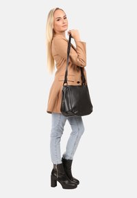 Zwarte leren handtas met een gladde textuur, voorzien van een enkele schouderband en een zijvak met rits, vastgehouden door een persoon in een lichtbruine jas.