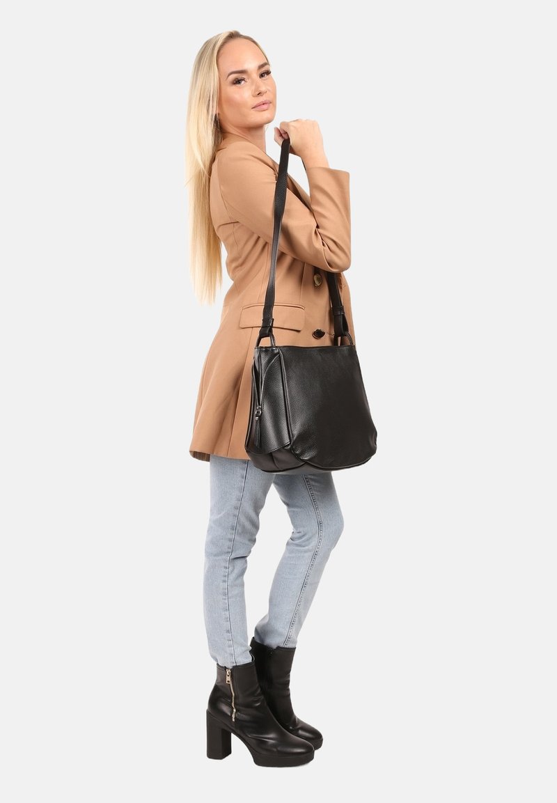 Zwarte leren handtas met een gladde textuur, voorzien van een enkele schouderband en een zijvak met rits, vastgehouden door een persoon in een lichtbruine jas.