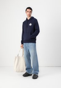 Felpa blu navy con tasca frontale e logo bianco, abbinata a jeans azzurri a gamba larga, scarpe nere e una borsa tote beige.