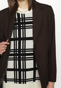 Blazer marron porté sur un pull à carreaux noir et blanc, présentant une texture côtelée. Le pull comporte des lignes horizontales et verticales audacieuses.