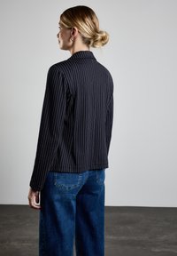 Svart och vitrandig blazer med en notch-krage, långa ärmar och rundad nederkant, parat med blå vida jeans, sedd från baksidan.