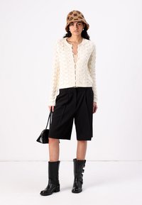 Kremfarget strikket cardigan med diamantmønster, svarte skråshorts, knehøye svarte støvler, og en brun bøttehatt med blomstermønster.