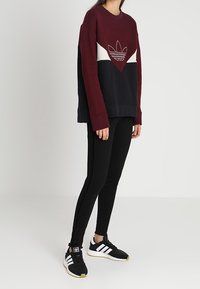 Osoba v bordo, bílé a černé mikině Adidas, černých legínách a černých teniskách Adidas s bílými pruhy na jednobarevném pozadí.