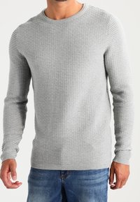 Homme portant un pull en maille texturée gris clair à manches longues et un jean bleu devant un fond uni.