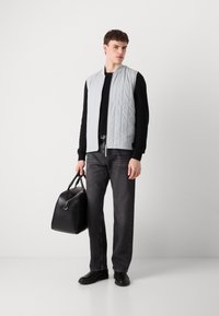 Colete acolchoado cinza claro sobre um suéter preto, combinado com jeans cinza escuro e sapatos pretos, segurando uma bolsa de couro preta. Design simples e funcional.