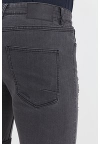 Jeans di denim nero con una superficie leggermente testurizzata, dotati di una tasca posteriore standard e di una targhetta di pelle con marchio sulla vita.