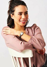 Silberne Metallarmbanduhr mit rechteckigem Zifferblatt. Frau trägt eine hellrosa strukturierte Bluse und sitzt mit verschränkten Armen auf einem weißen Stuhl.