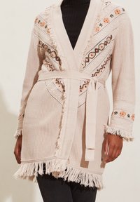Beige knitted cardigan featuring embroidered floral patterns, fringe detailing along the edges, and a tied belt at the waist.