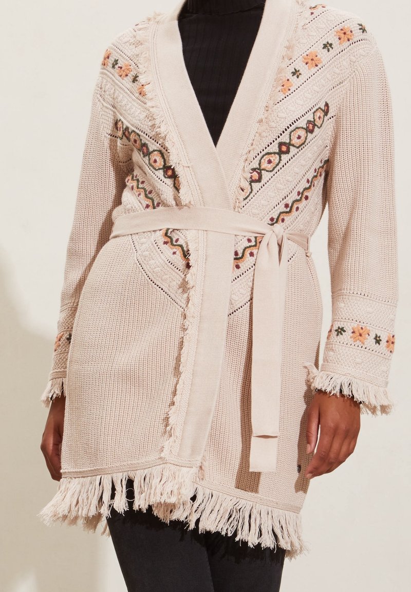 Beige knitted cardigan featuring embroidered floral patterns, fringe detailing along the edges, and a tied belt at the waist.