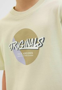Svijetlozeleni pamučni majica s okruglim grafičkim prikazom u bež i ljubičastoj boji s natpisima "ORIGINALS" i "JACK AND JONES" ispod.