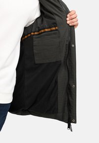 Mørkegrøn jakke indeni med et struktureret mønster, der har en orange stribet detalje og en lomme på venstre side. Glat stof, lynlås lukning.