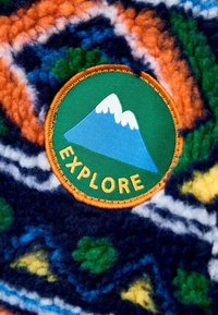 Un tessuto in pile colorato presenta un motivo arancione e blu marino con accenti verdi e gialli. Una toppa circolare mostra montagne e la parola "ESPLORA".