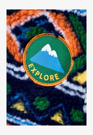 Barvna flis tkanina ima vzorec v oranžni in mornarsko modri barvi s zelenimi in rumenimi poudarki. Okrogla zaklopeka prikazuje gore in besedo "RAZISKUJ."