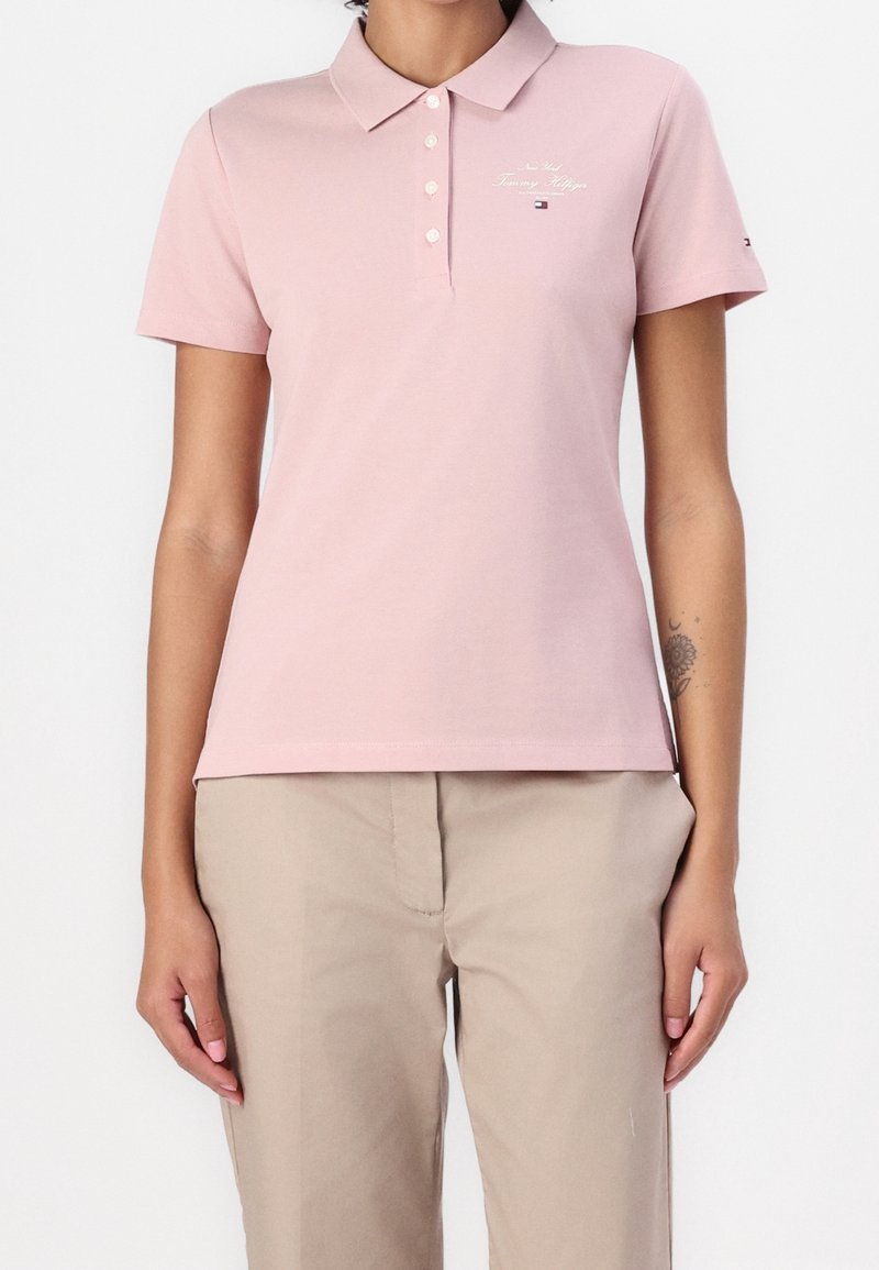 Femme portant un polo rose clair à manches courtes et un pantalon beige, debout avec les bras détendus le long du corps, sur un fond blanc.