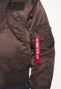 Hnědá bomber bunda Alpha Industries s kapsou na zip na rukávu a červeným textilním štítkem "Remove Before Flight".