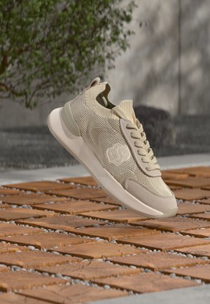 Baskets en mesh beige clair avec lacets flottant au-dessus de carreaux en bois à l'extérieur, avec une plante verte et un mur en béton en arrière-plan.