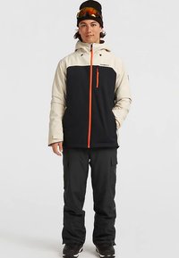 Giovane con una giacca da sci nera e beige, pantaloni da sci neri, un berretto nero e occhiali da sci con lenti arancioni sulla testa.