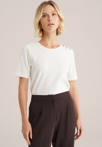 Femeie cu părul blond scurt, purtând un top alb cu nasturi pe umăr și pantaloni maro, stând pe un fundal deschis.