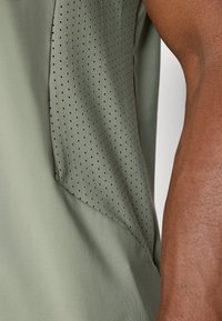 Mouwloos topje van een licht olijfgroene stof, met een gladde textuur en geperforeerde donkerder groene accenten langs een kant.