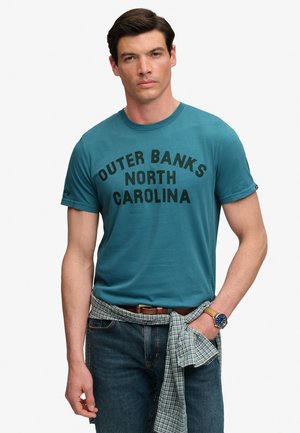Mężczyzna noszący turkusową koszulę z tekstem "Outer Banks North Carolina", jeansy, koszulę w kratę zawiązaną w pasie oraz zegarek na lewej ręce, stojący na białym tle.