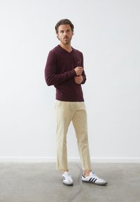 Bordeaux v-hals trui, crèmekleurige chinos en grijs-witte sneakers met zwarte strepen. Glad, snug-fitting stof; eenvoudig ontwerp; geen zichtbare patronen.
