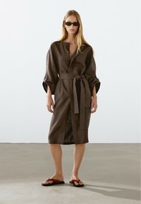 Robe en lin marron avec un devant à boutons, des manches larges et une taille nouée. Le modèle porte des tongs marron et rouge, posant sur un sol neutre.