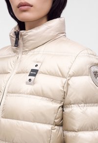 Femme portant une doudoune beige clair avec col montant, fermeture éclair sur le devant, et des étiquettes du logo Blauer sur la poitrine et le haut de la manche.
