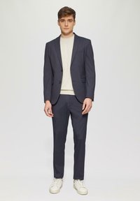 Veste de costume bleu marine avec revers en angle, associée à un léger pull et un pantalon assorti. Des baskets blanches ajoutent une touche décontractée.