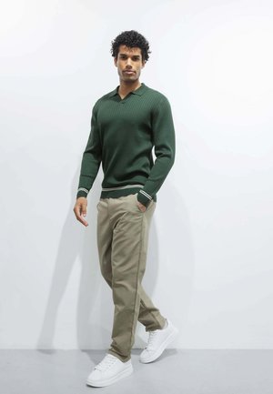 Homme portant un pull côtelé vert foncé, pantalon beige, baskets blanches, debout contre un mur blanc avec une main dans la poche et marchant en avant.