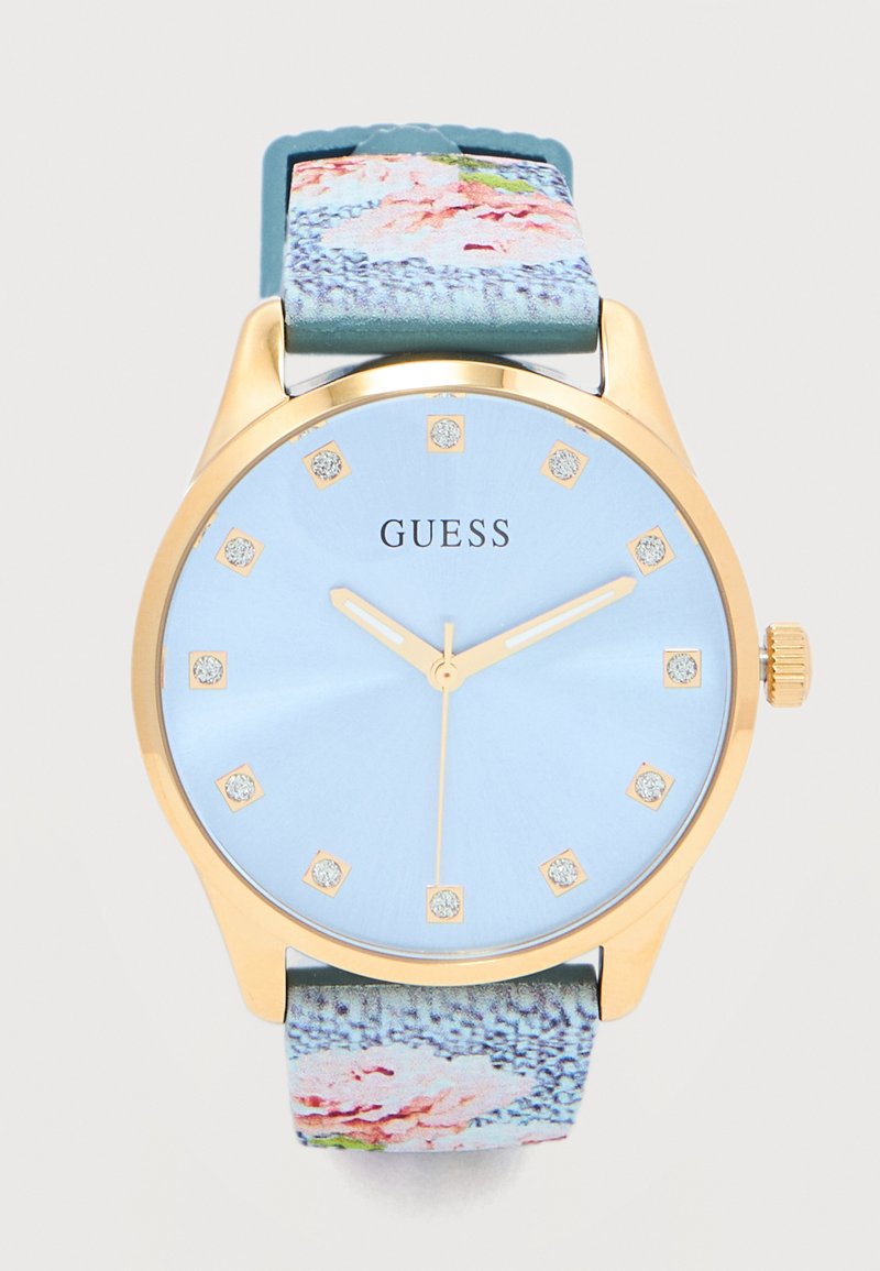 Στρογγυλό ρολόι χειρός Guess με επιχρυσωμένο τόνο, ανοιχτό μπλε καντράν, κρυστάλλινους δείκτες ώρας, χρυσά λεπτά και λουρί από ύφασμα με ανθισμένο σχέδιο.