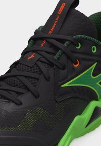 Αθλητικό παπούτσι σε μαύρο και πράσινο με υφασμάτινο δίχτυ, μαύρα κορδόνια, φωτεινή πράσινη σόλα και πορτοκαλί λεπτομέρειες στα οπές για κορδόνια και τη σήμανση.
