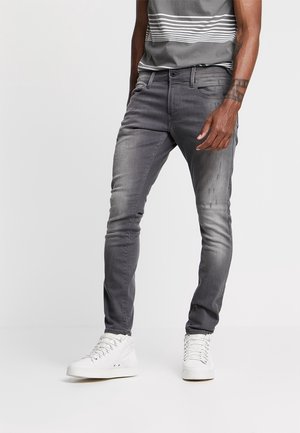 Homme portant un jean slim gris délavé, un t-shirt rayé gris, des baskets montantes blanches et un tatouage au poignet, debout devant un fond blanc uni.
