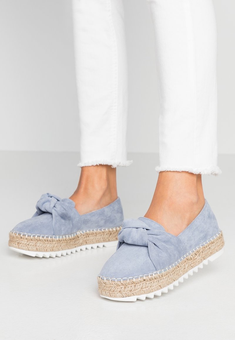 Espadrilles en suède bleu clair avec un nœud, semelle intermédiaire en jute et semelle en caoutchouc blanche. Un jean blanc avec un ourlet effiloché est porté avec.