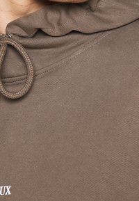 Acercamiento de la tela marrón de una sudadera con capucha, con bucle de cordón y texto blanco parcial en el pecho.