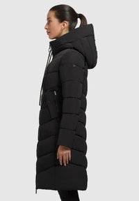 Abrigo negro largo tipo plumón con acolchado en forma de chevron, capucha, cordón ajustable y bolsillos laterales con cremallera. Textura suave, cierre con cremallera.