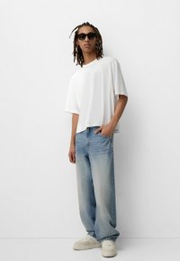 Weißes, kurzärmliges T-Shirt in lässigem Schnitt; hellblaue, weite Jeans; klobige weiße Sneakers; schwarze Sonnenbrille; glatte Texturen.