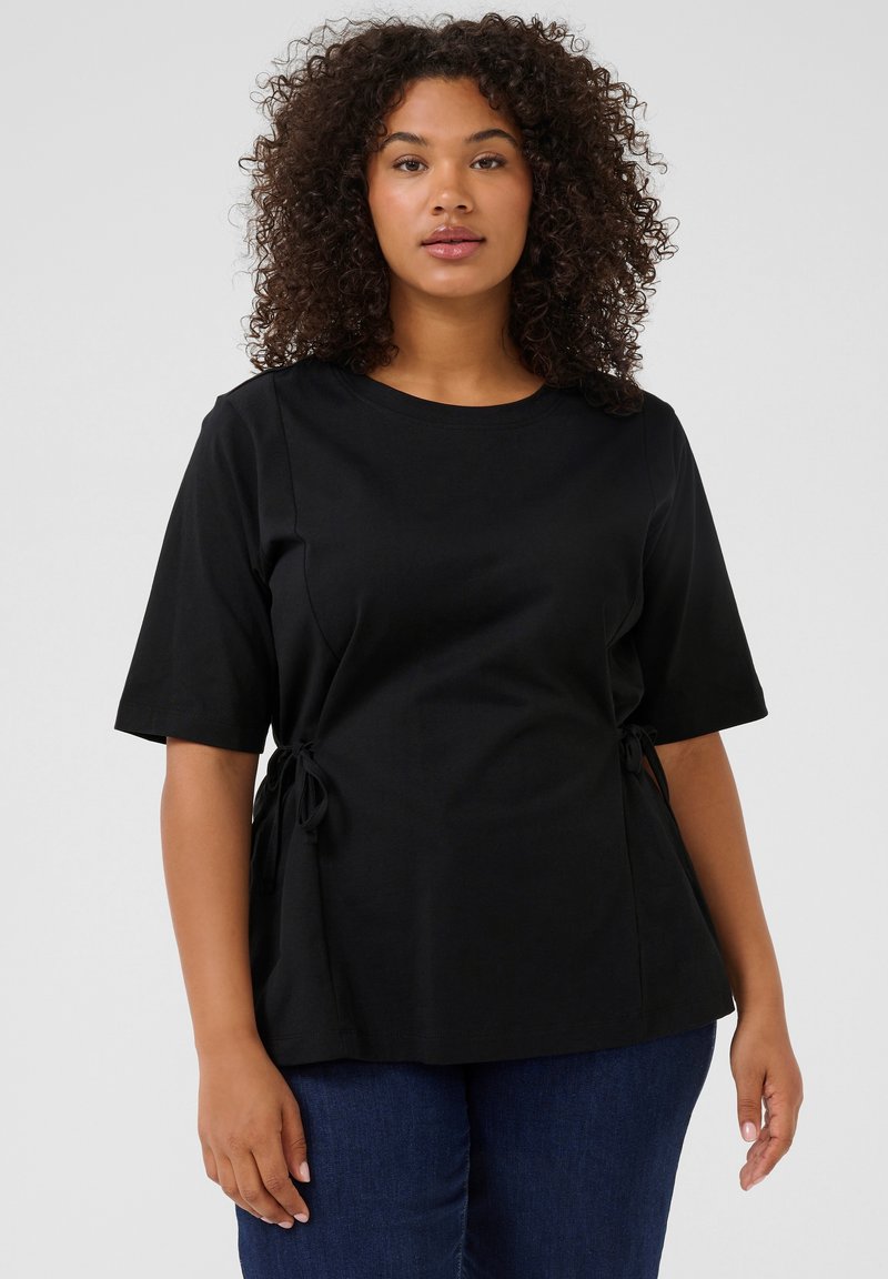 Camiseta negra de manga corta con cuello redondo, con detalles de lazo en el costado, fabricada en una tela suave. Combinada con jeans de mezclilla azules.